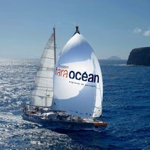 a research sailboat with white sails inscribed with "tara ocean" traverses a body of water with small, rocky islands in the background