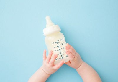Infant hands holding bottle of milk on light blue floor background.
