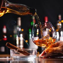 a person pours whiskey into a glass held by another person, with a smoking cigar in a tray in front