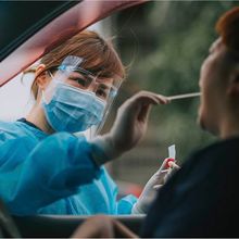 Woman in face shield and blue gown taking cotton swab of patient's mouth while patient sits inside of car