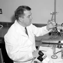 Man in lab coat sitting at a lab bench looking at small, stoppered beaker.